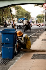 A person wearing a yellow uniform and orange hat is crouching beside a blue trash bin on a city sidewalk. A long broom is laying on the ground next to them. In the background, there is a busy street with vehicles and a bridge overhead. Trees line the side of the road amidst the urban setting.