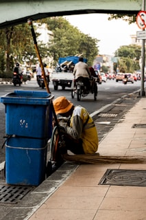 A person wearing a yellow uniform and orange hat is crouching beside a blue trash bin on a city sidewalk. A long broom is laying on the ground next to them. In the background, there is a busy street with vehicles and a bridge overhead. Trees line the side of the road amidst the urban setting.