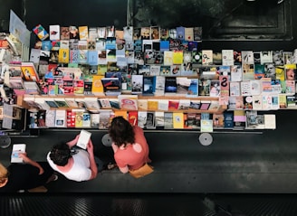 Aerial view of a table filled with a wide variety of books arranged neatly. Three people stand in front browsing through the selection. The books have colorful covers, and the scene is indoors with a modern, dark background.