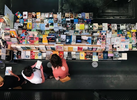 Aerial view of a table filled with a wide variety of books arranged neatly. Three people stand in front browsing through the selection. The books have colorful covers, and the scene is indoors with a modern, dark background.