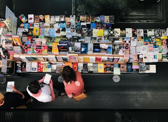 Aerial view of a table filled with a wide variety of books arranged neatly. Three people stand in front browsing through the selection. The books have colorful covers, and the scene is indoors with a modern, dark background.