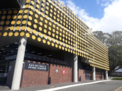 The image features a building with an exterior facade covered in a pattern of large yellow circular discs. The structure has a modern design with grey concrete and brick elements. There is a sign indicating 'Race Day Stall & Owners Ticketing', and the setting appears to be an outdoor venue. The scene is bright and sunny with clear skies and some trees visible in the background.