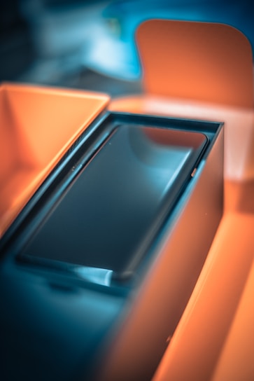 A close-up view of a sleek black electronic device, possibly a smartphone, placed inside an open orange box. The focus is on the device with its glossy screen, and the background is blurred, highlighting shades of blue and orange.