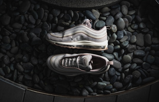 Two sneakers are artfully placed on a bed of smooth, dark pebbles. The shoes have a sleek, modern design with wavy patterns and a translucent air cushion sole. The lighting casts soft shadows, highlighting the texture of both the sneakers and the stones.