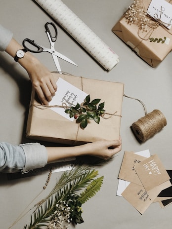 A person is wrapping a gift in brown paper and tying it with twine. The setup includes decorative elements like green leaves and small berries. There are scissors, a roll of wrapping paper, and cards with handwritten messages. Additional decorative foliage and sprigs are spread around, creating a rustic and handcrafted ambiance.