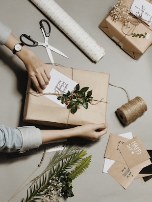 A person is wrapping a gift in brown paper and tying it with twine. The setup includes decorative elements like green leaves and small berries. There are scissors, a roll of wrapping paper, and cards with handwritten messages. Additional decorative foliage and sprigs are spread around, creating a rustic and handcrafted ambiance.