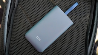 A sleek, matte-finished power bank with the number 5100 on it lies on a textured dark material. A blue strap is attached to the power bank.