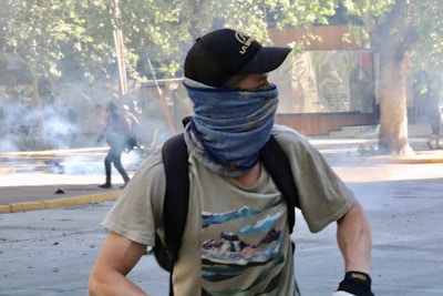 A person wearing a cap and a bandana over their face, possibly engaged in a protest or demonstration. Smoke billows in the background, along with some trees and urban structures.