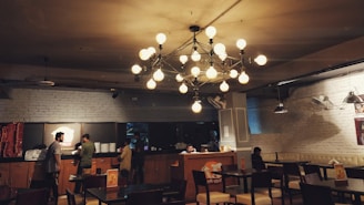 A cozy restaurant interior with dim lighting, featuring a large, modern chandelier with spherical bulbs. There are several tables and chairs arranged throughout the room. On the left, a counter with plates and red packaging is visible, along with a few people standing and interacting. The walls are brick, painted white, and there are spotlights mounted on the ceiling.
