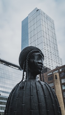 A large, dark-colored sculpture of a figure with braided hair is in the foreground. The sculpture appears to be made of metal and has a detailed, textured surface. Behind the sculpture, there are modern skyscrapers and buildings with glass facades, reflecting the cloudy sky above.