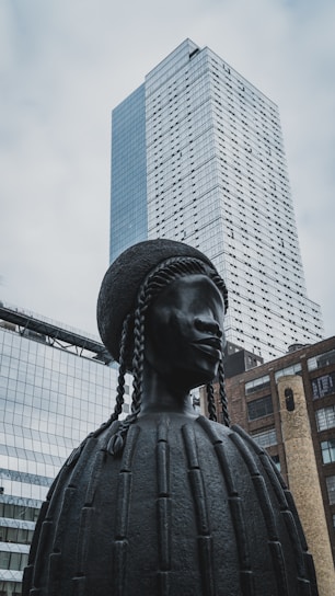 A large, dark-colored sculpture of a figure with braided hair is in the foreground. The sculpture appears to be made of metal and has a detailed, textured surface. Behind the sculpture, there are modern skyscrapers and buildings with glass facades, reflecting the cloudy sky above.