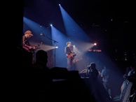 Musicians perform on a dimly lit stage with spotlights casting beams of light. The main performer plays a guitar and sings into a microphone, while another musician is seen playing a guitar in the background. Audience members are visible in silhouette, adding to the dynamic atmosphere of the live concert setting.