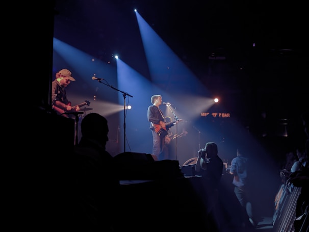 Musicians perform on a dimly lit stage with spotlights casting beams of light. The main performer plays a guitar and sings into a microphone, while another musician is seen playing a guitar in the background. Audience members are visible in silhouette, adding to the dynamic atmosphere of the live concert setting.