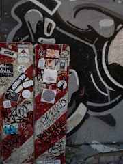 A wall features a vibrant collection of graffiti and street art. It includes a large black and white abstract design in the background. In the foreground, a panel is covered with various colorful stickers and tags, reflecting urban art culture.