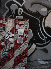 A wall features a vibrant collection of graffiti and street art. It includes a large black and white abstract design in the background. In the foreground, a panel is covered with various colorful stickers and tags, reflecting urban art culture.