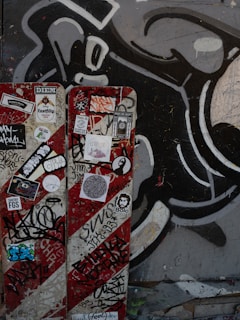 A wall features a vibrant collection of graffiti and street art. It includes a large black and white abstract design in the background. In the foreground, a panel is covered with various colorful stickers and tags, reflecting urban art culture.