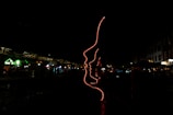 A dark environment featuring an artistic installation made of red neon lights outlining several human profiles. The setting appears to be an outdoor area, possibly near shops or cafes, indicated by faintly visible structures with subtle lighting in the background.