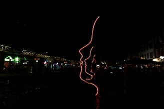 A dark environment featuring an artistic installation made of red neon lights outlining several human profiles. The setting appears to be an outdoor area, possibly near shops or cafes, indicated by faintly visible structures with subtle lighting in the background.