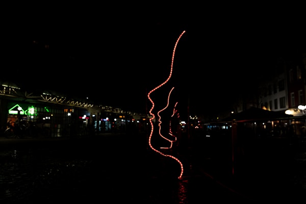 A dark environment featuring an artistic installation made of red neon lights outlining several human profiles. The setting appears to be an outdoor area, possibly near shops or cafes, indicated by faintly visible structures with subtle lighting in the background.