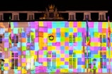 A building facade illuminated with a colorful grid pattern of squares in various hues. The bright projection casts an array of colors across the architectural details, including windows and decorative elements. Flags are visible on the sides, adding to the vibrant ambiance.