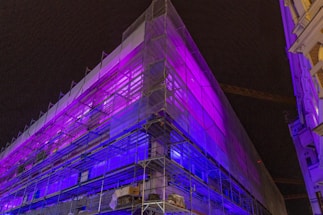 A building under construction or renovation is wrapped in a semi-transparent covering, illuminated by vibrant purple and blue lights against a dark night sky. Scaffolding frames the structure, and there are two adjacent buildings on either side.