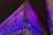A building under construction or renovation is wrapped in a semi-transparent covering, illuminated by vibrant purple and blue lights against a dark night sky. Scaffolding frames the structure, and there are two adjacent buildings on either side.