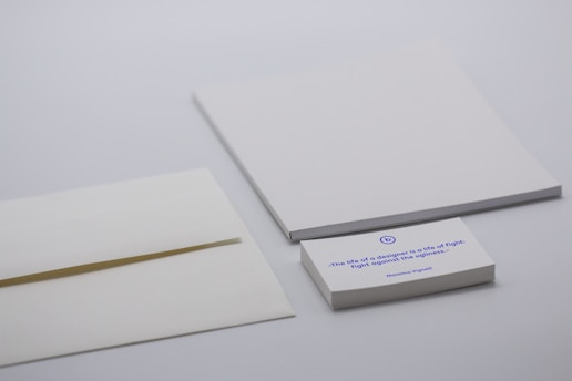 A minimalist desk setup with a focus on stationery items including two blank white sheets, an envelope, and a stack of cards. The card on the top features a printed quote in blue ink. The background is a neutral light gray.