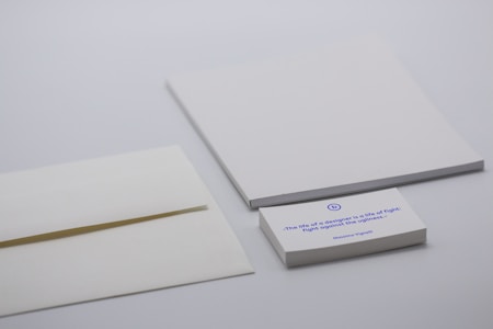 A minimalist desk setup with a focus on stationery items including two blank white sheets, an envelope, and a stack of cards. The card on the top features a printed quote in blue ink. The background is a neutral light gray.