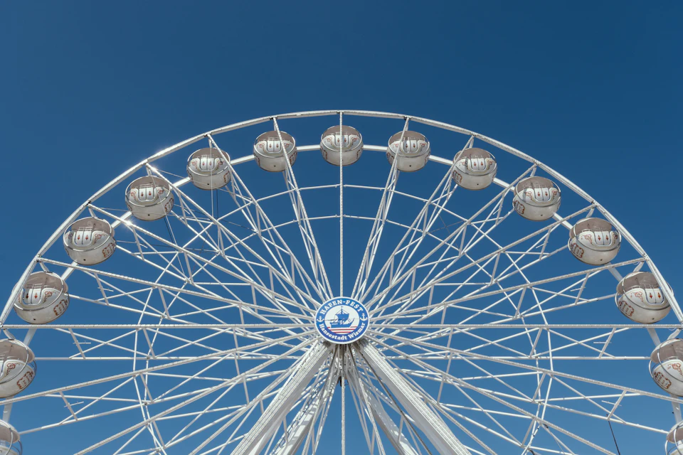 The image shows a large white Ferris wheel with 12 evenly spaced round gondolas, each adorned with a blue and white logo on top. The gondolas are arranged in two rows, with 4 at the front and 8 at the back, creating a symmetrical pattern. The Ferris wheel is set against a clear blue sky, which serves as a perfect backdrop for this iconic amusement park attraction.