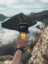 A hand holding a small camera is positioned in front of a scenic landscape with mountains, a body of water, and clouds reflecting on the water's surface. The camera's display shows the same landscape, capturing the picturesque view.