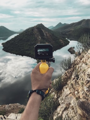 A hand holding a small camera is positioned in front of a scenic landscape with mountains, a body of water, and clouds reflecting on the water's surface. The camera's display shows the same landscape, capturing the picturesque view.