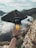 A hand holding a small camera is positioned in front of a scenic landscape with mountains, a body of water, and clouds reflecting on the water's surface. The camera's display shows the same landscape, capturing the picturesque view.