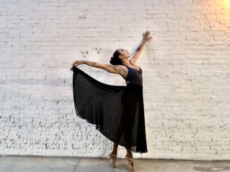 A dancer poses gracefully against a white brick wall. The dancer is wearing a dark sheer skirt and pointe shoes, with arms extended elegantly, creating a sense of movement and balance.
