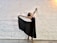 A dancer poses gracefully against a white brick wall. The dancer is wearing a dark sheer skirt and pointe shoes, with arms extended elegantly, creating a sense of movement and balance.