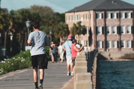 A scenic waterfront walkway with people walking and jogging. Tall buildings and palm trees are in the background, and the walkway is bordered by a body of water on one side. The atmosphere appears relaxed and casual, with people enjoying a sunny day.