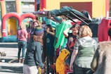 A group of people gathered around the back of a car with its trunk open, participating in an outdoor event. Some individuals are dressed in casual clothing, while a child is wearing a yellow Pikachu costume. In the background, inflatable structures suggest a festive or family-oriented gathering.