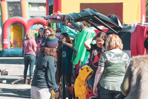 A group of people gathered around the back of a car with its trunk open, participating in an outdoor event. Some individuals are dressed in casual clothing, while a child is wearing a yellow Pikachu costume. In the background, inflatable structures suggest a festive or family-oriented gathering.