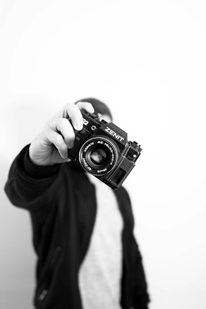 A person holds a vintage Zenit camera close to the lens, with their hand in focus while their face and body are blurred in the background. The image is in black and white, giving it a classic and timeless feel.