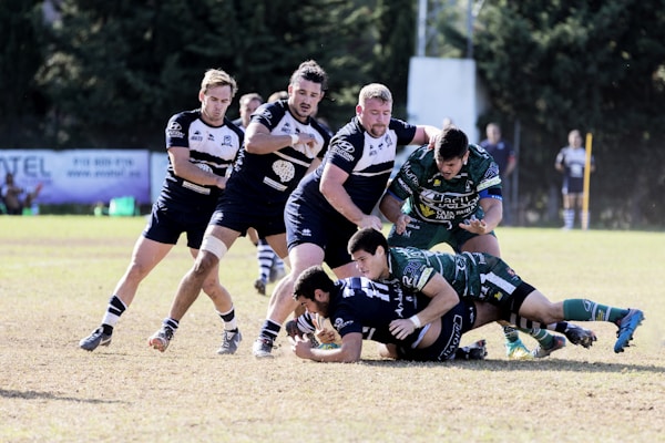 A group of rugby players engaged in a dynamic play on the field. Two players in green jerseys are tackling an opposing player who is holding the ball, while teammates in dark blue jerseys are moving in to support. The grassy field and blurred spectators in the background suggest an outdoor sports environment.
