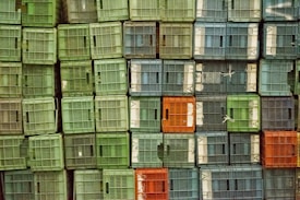 A stack of plastic crates organized in a grid-like pattern with various shades of green, blue, and a few orange crates. The crates have slatted sides, allowing for ventilation, and are tightly packed together. Some crates are secured with white ties.