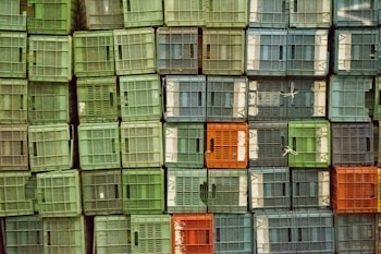 A stack of plastic crates organized in a grid-like pattern with various shades of green, blue, and a few orange crates. The crates have slatted sides, allowing for ventilation, and are tightly packed together. Some crates are secured with white ties.