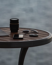 On a round wooden table, there is a camera lens, a pair of sunglasses, a closed notebook, and a smartphone. The background shows a blurred view of water, suggesting an outdoor setting.