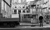 A construction site with an unfinished building surrounded by scaffolding and protective barriers. A person wearing a hard hat stands observing the progress, next to a truck bearing the signage of an architecture firm. Stacks of materials and construction equipment are visible in the foreground.