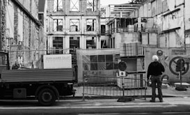 A construction site with an unfinished building surrounded by scaffolding and protective barriers. A person wearing a hard hat stands observing the progress, next to a truck bearing the signage of an architecture firm. Stacks of materials and construction equipment are visible in the foreground.