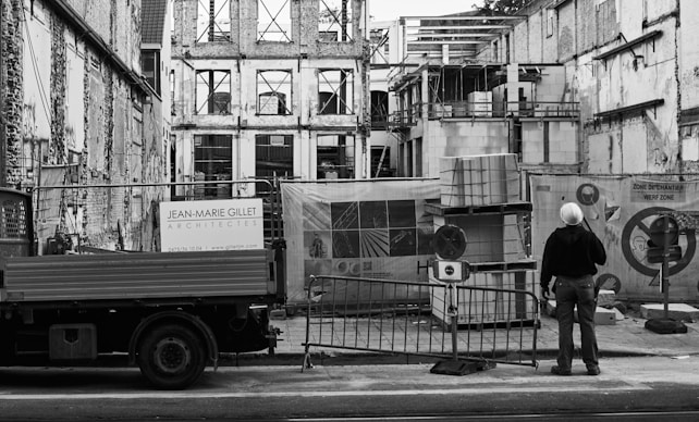 A construction site with an unfinished building surrounded by scaffolding and protective barriers. A person wearing a hard hat stands observing the progress, next to a truck bearing the signage of an architecture firm. Stacks of materials and construction equipment are visible in the foreground.