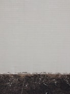 A simple tiled wall with small beige tiles that seem slightly aged, meeting a dark, worn concrete floor at the bottom. Some white marks and debris are visible on the floor.