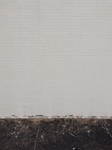 A simple tiled wall with small beige tiles that seem slightly aged, meeting a dark, worn concrete floor at the bottom. Some white marks and debris are visible on the floor.