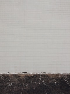 A simple tiled wall with small beige tiles that seem slightly aged, meeting a dark, worn concrete floor at the bottom. Some white marks and debris are visible on the floor.