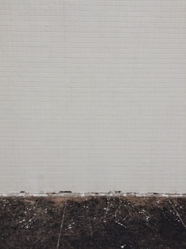 A simple tiled wall with small beige tiles that seem slightly aged, meeting a dark, worn concrete floor at the bottom. Some white marks and debris are visible on the floor.