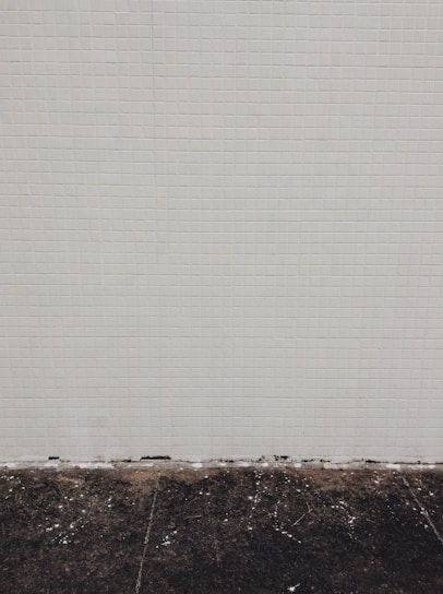 A simple tiled wall with small beige tiles that seem slightly aged, meeting a dark, worn concrete floor at the bottom. Some white marks and debris are visible on the floor.